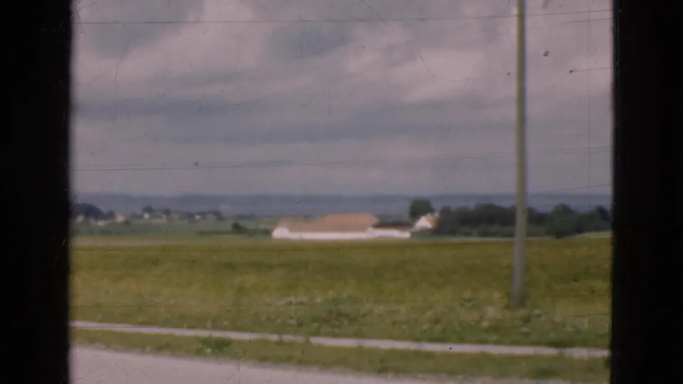 1975 Denmark home-movie frame of a rural field and roadside, with a distant low building, utility pole, hills, and visible 8mm wear.