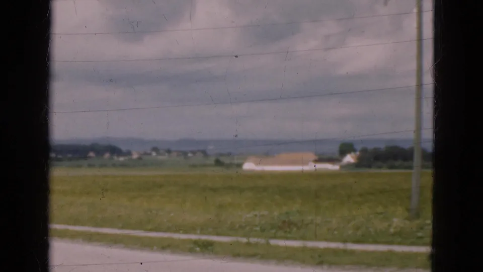 1975 Denmark countryside frame showing an open field, pale building with tan roof, roadside edge, utility pole, and dark window border.