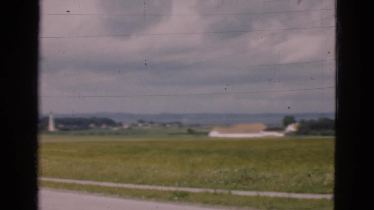 1975 Danish 8mm frame of green farmland with a distant white building and lighthouse-like tower, partially framed by dark window edges.