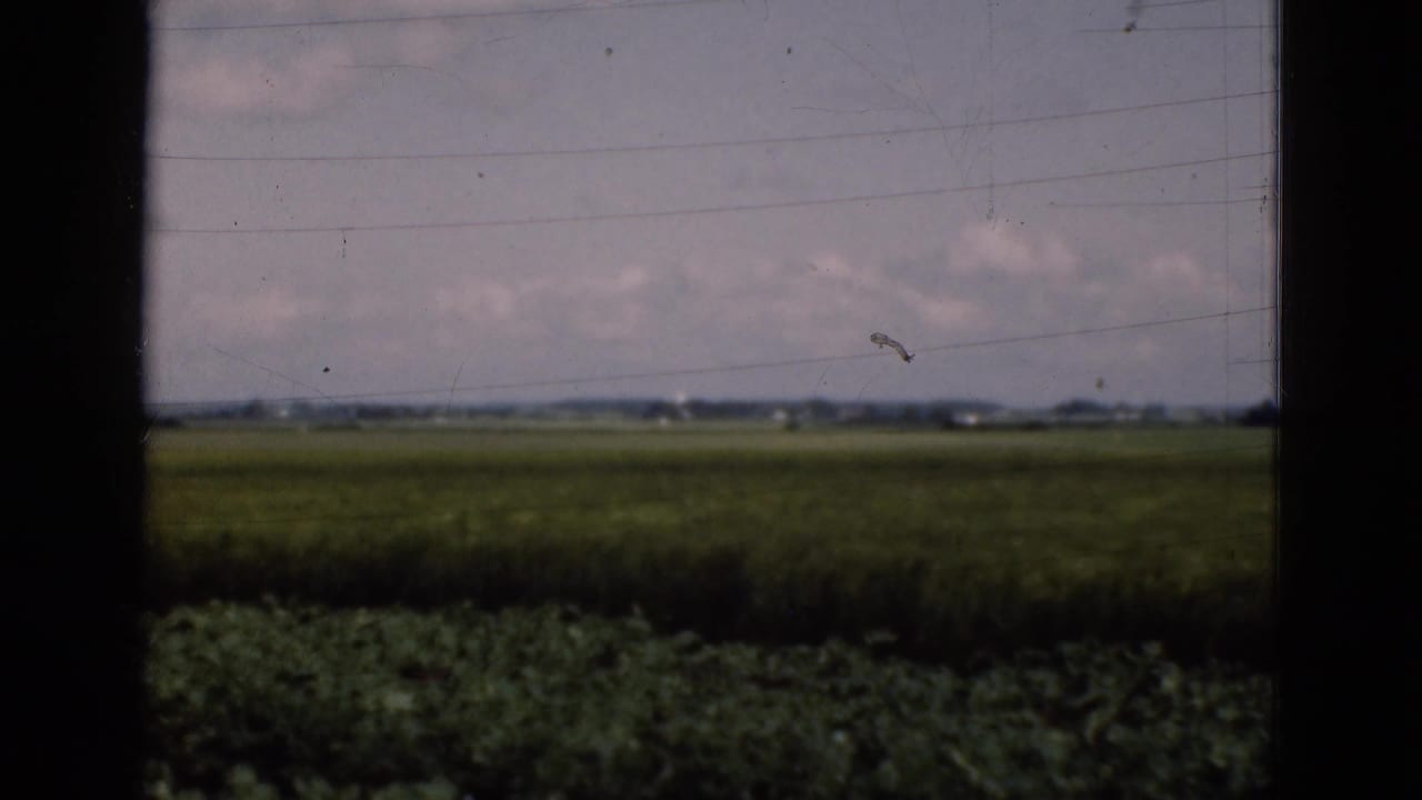 Rural Danish farmland in a 1975 8mm frame, seen through a vehicle or window with dark vertical borders and distant horizon buildings.