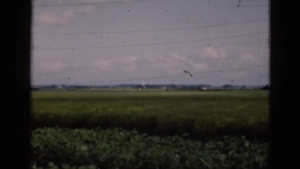 Rural Danish farmland in a 1975 8mm frame, seen through a vehicle or window with dark vertical borders and distant horizon buildings.