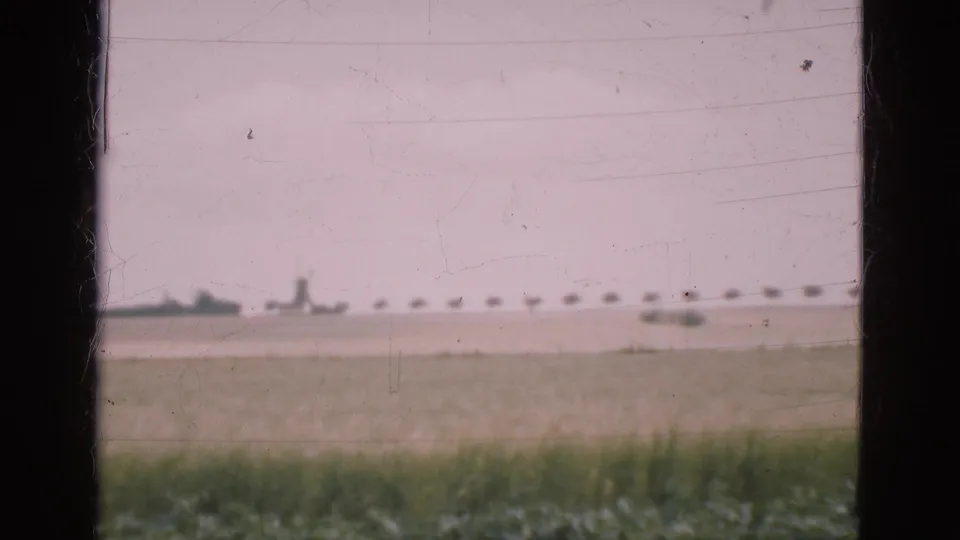 A passing rural view shows a green field, pale sky, and distant shapes on the horizon, partly blocked by a window frame.