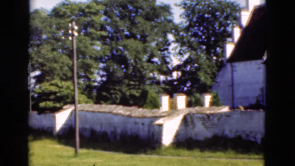 1975 Denmark exterior with a low whitewashed wall, grassy foreground, utility pole, trees, and a tall white building at right.