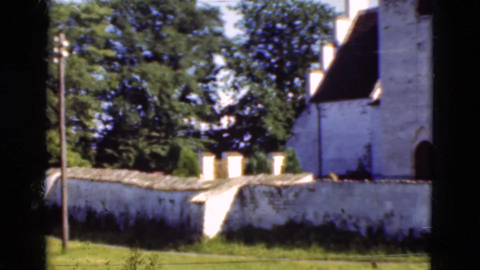 1975 Denmark 8mm frame showing a whitewashed building, dark roof, arched doorway, low wall, grass, and trees.