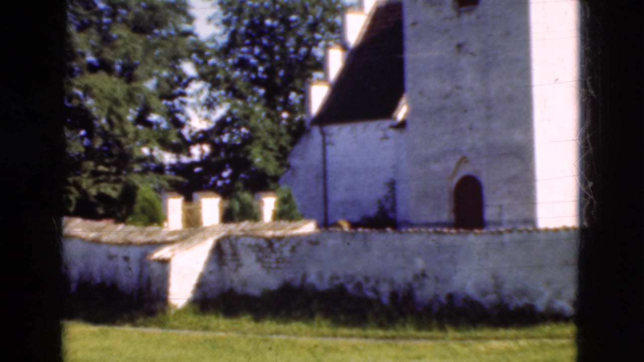 1975 Denmark 8mm frame showing a white church exterior, low wall, trees, grass, and dark film-edge vignetting.