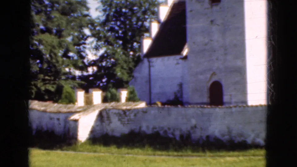 1975 Denmark 8mm frame showing a white church exterior, low wall, trees, grass, and dark film-edge vignetting.