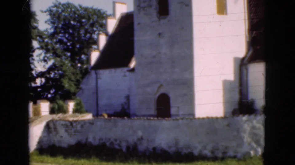 1975 Denmark 8mm frame showing a closer view of the white church tower, dark arched door, low wall, roof, and trees.