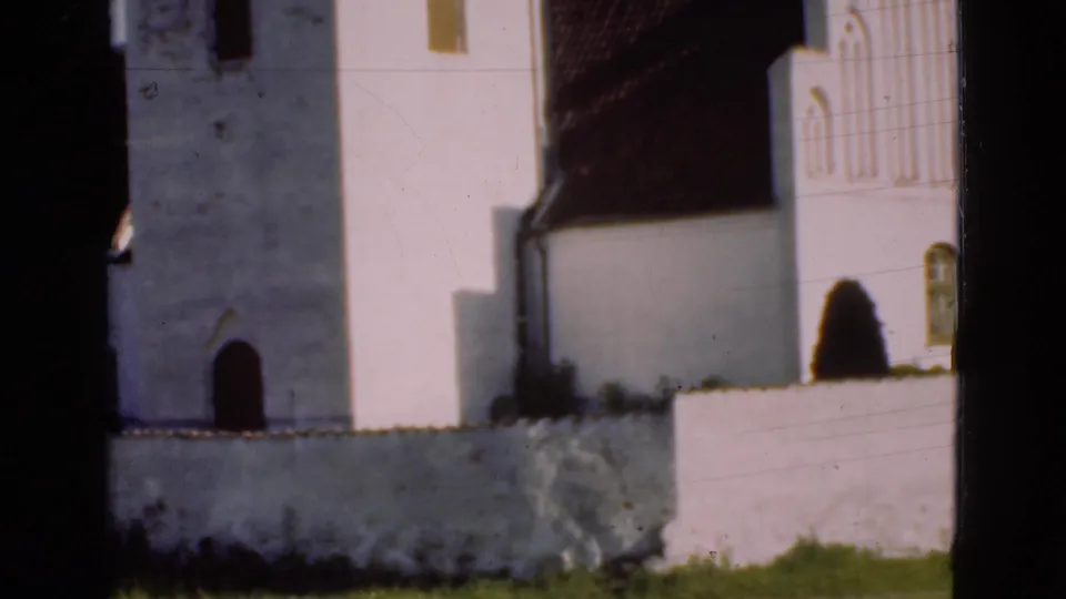 1975 Denmark 8mm frame showing the white church tower and adjacent building mass behind a low wall, with grass and soft film exposure.