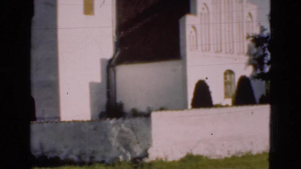 1975 Denmark 8mm frame showing the white church facade, tower edge, arched architectural details, low wall, grass, and dark shrubs.