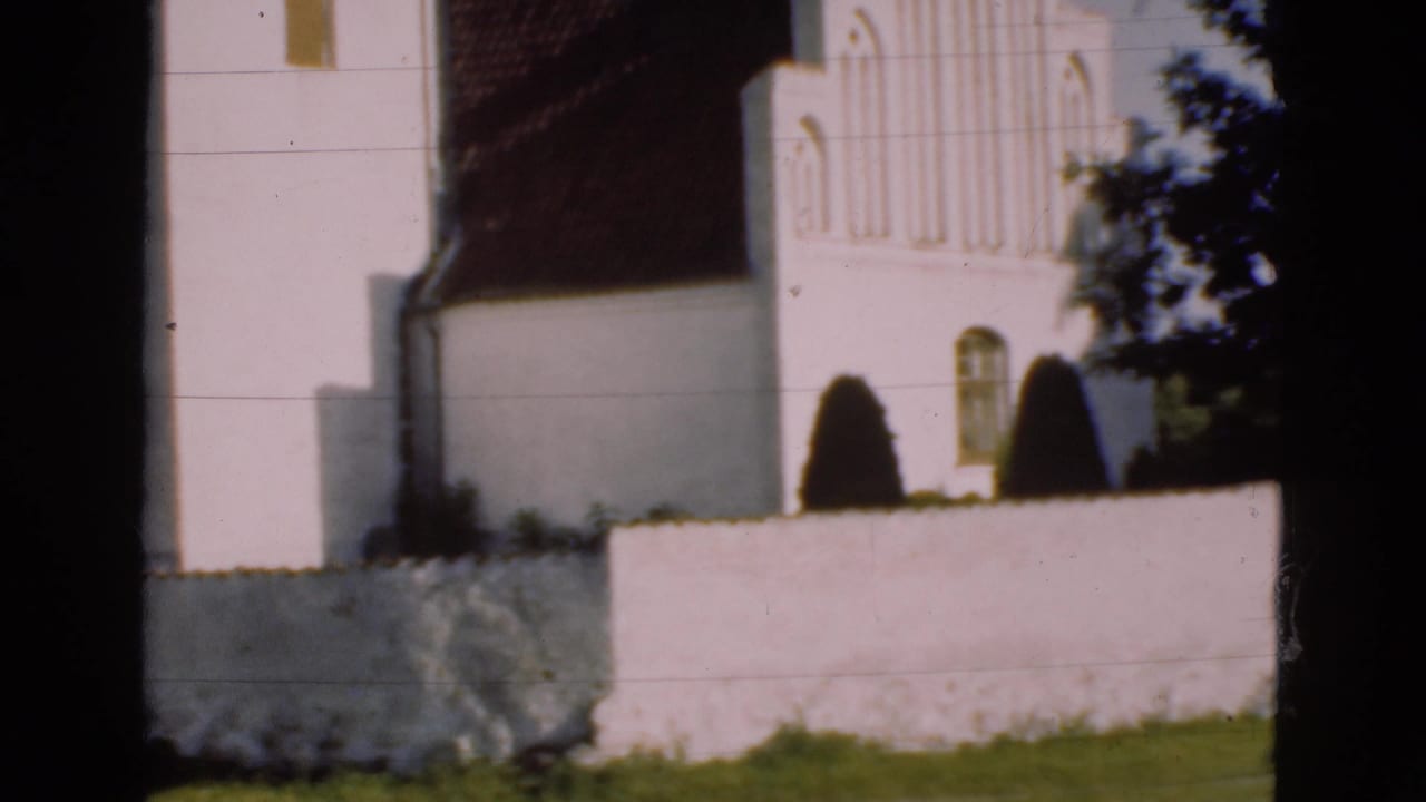 White church facade behind a low wall with shrubs and tree shadow.