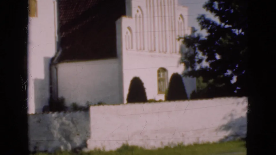 1975 Denmark 8mm frame showing the church from across a low wall, with Gothic-style facade details, shrubs, grass, and soft film exposure.