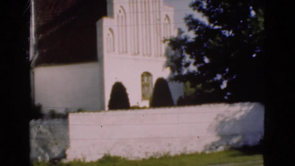 1975 Denmark 8mm frame of a white church exterior behind a low wall, with shrubs near the facade and dark tree branches obscuring part of the view.
