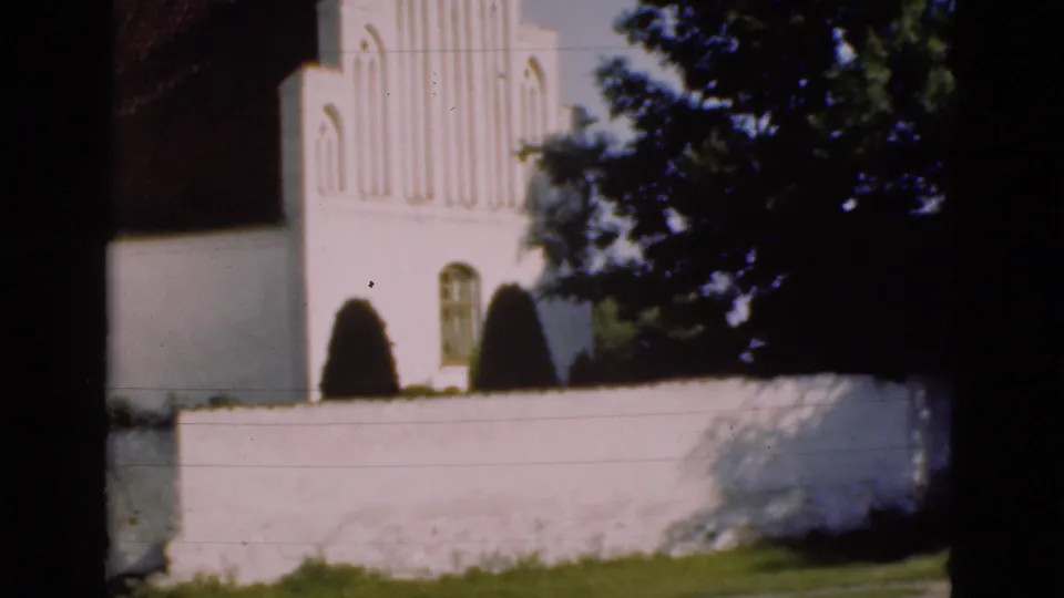 1975 Denmark 8mm frame showing a white church exterior partly hidden by a wall, shrubs, trees, and heavy shadow.