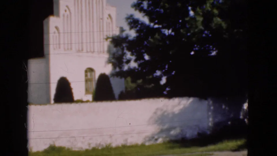 White church exterior in Denmark, filmed on 8mm in 1975, seen across a wall with trees obscuring part of the facade.