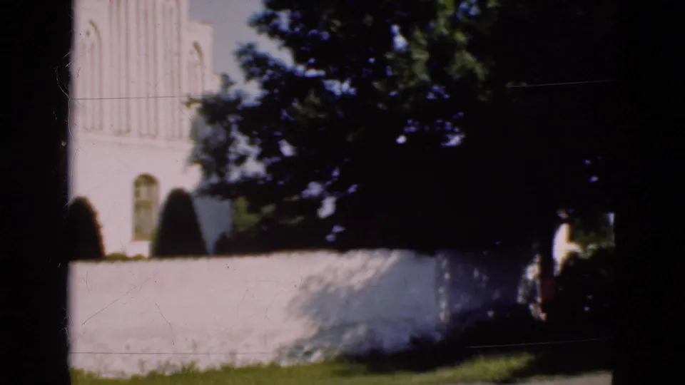 Soft 8mm frame of a Danish church exterior, mostly obscured by trees, wall, and dark foreground shadow.