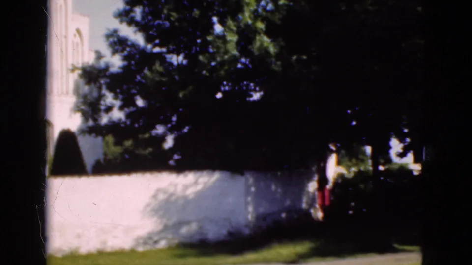 1975 Denmark 8mm frame of a white church wall and trees, with distant passersby briefly visible near the right side.