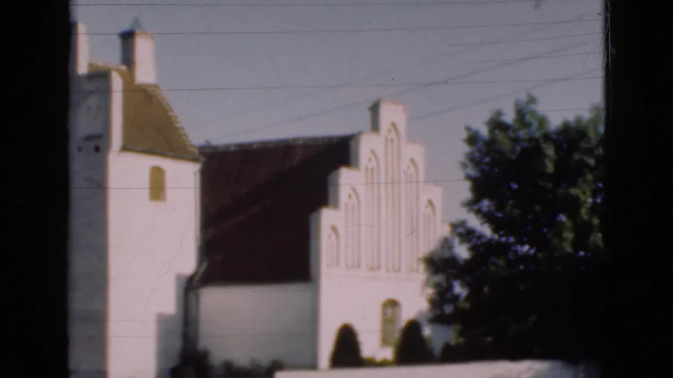 1975 Denmark 8mm frame showing the church side and stepped gable with a dark roof, leafy tree, low wall, and soft film grain.