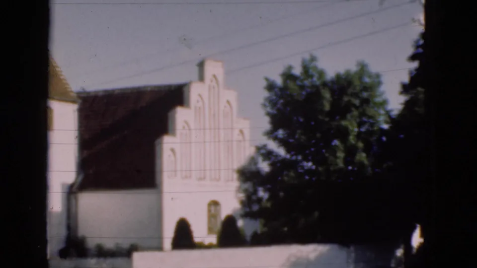 1975 Denmark 8mm frame with the white church gable and roof partly hidden by a large tree, low wall, and dark frame border.
