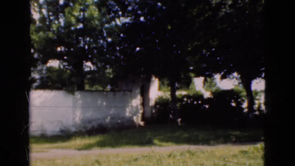 1975 Denmark 8mm frame looking past a white wall and trees over grass and a path as the camera pans away from the building.