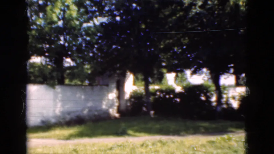 1975 Denmark 8mm frame showing leafy trees, a white boundary wall, grass, and a narrow path on the property edge.