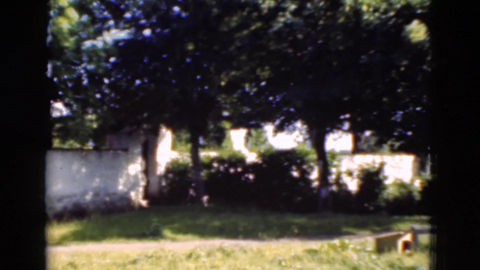 1975 Denmark 8mm frame of a white wall, shaded trees, grass, path, and a small low object near the lower right edge.