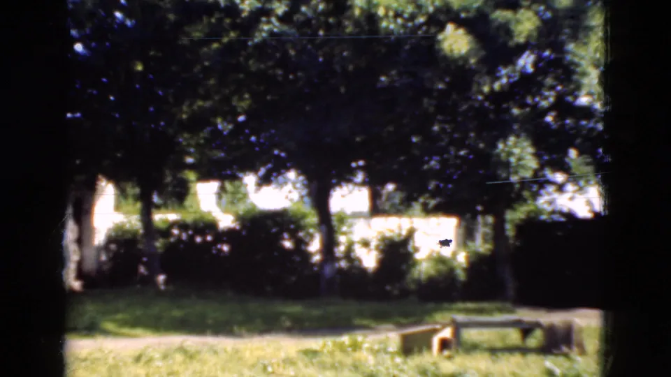 1975 Danish home-movie frame showing a leafy garden with shrubs, lawn, a simple bench, and white residential walls in the distance.