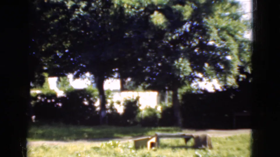 Soft 8mm frame from Denmark in 1975 showing a green garden, shrubs, white house wall, wooden bench, and shaded path area.