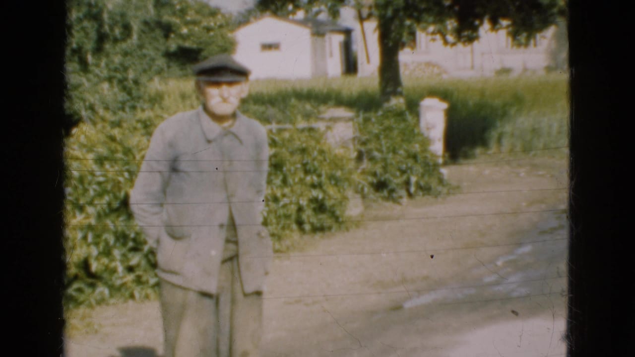 Elderly man in a flat cap standing in a rural driveway near hedges and a white building.