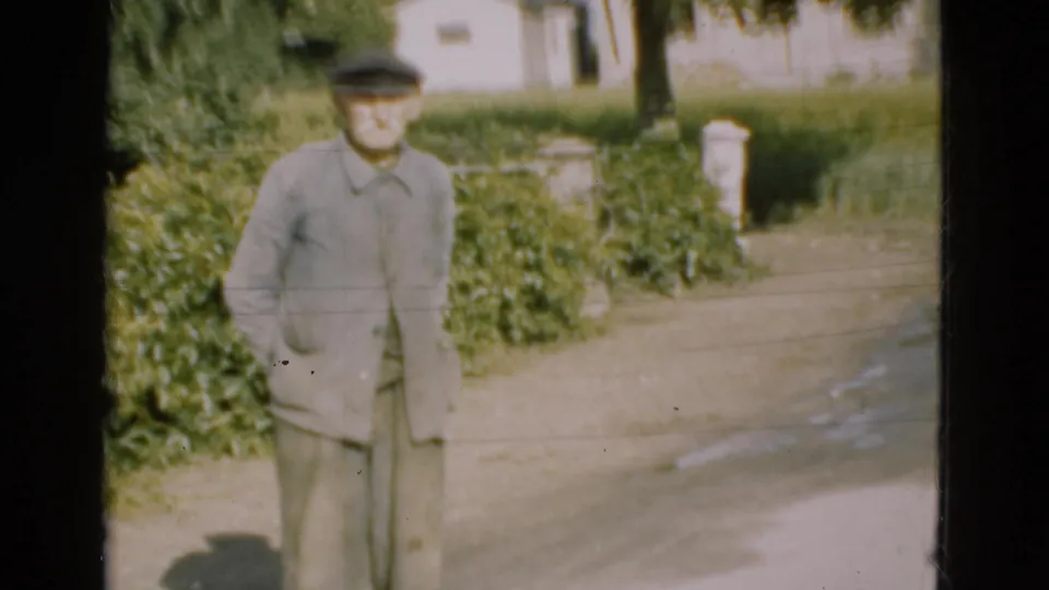 1975 Denmark home-movie frame showing an elderly man in light clothing and a flat cap beside a rural driveway and hedges.