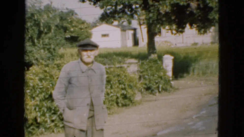 1975 Denmark home-movie frame of an elderly man posing near hedges, a driveway, trees, and a white rural building.