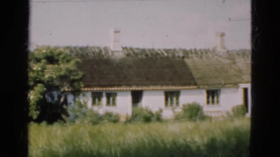 1975 Denmark 8mm frame showing a white thatched cottage across tall grass, partly framed by dark vertical window edges.