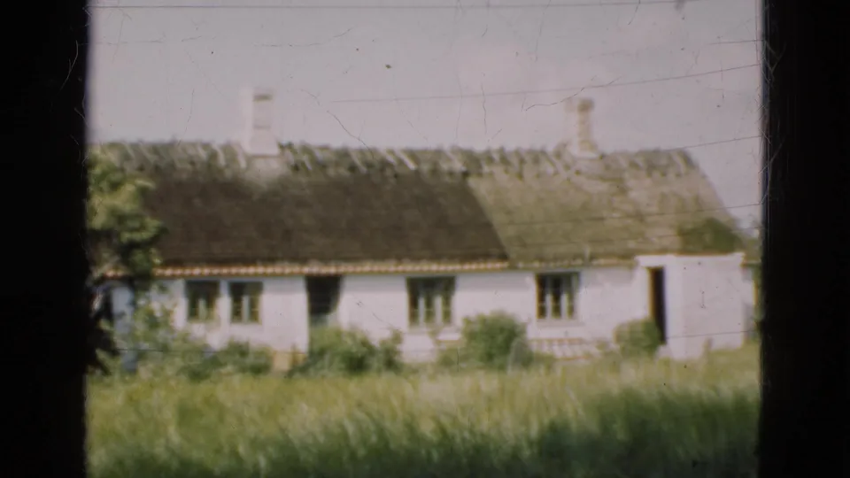 1975 Denmark 8mm frame of a traditional white cottage with a thatched roof, chimneys and tall grass in the foreground.