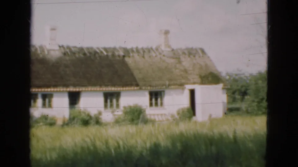 1975 Denmark 8mm frame showing the cottage shifted closer in view, with tall grass below and a dark window-frame edge at left.