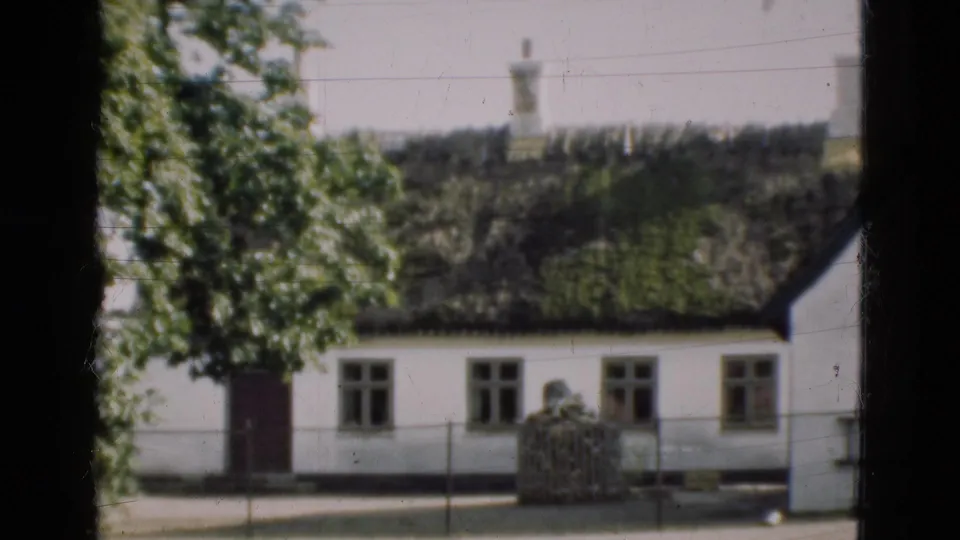 1975 Denmark 8mm frame showing another angle of the white thatched house with a tree, doorway, windows and a low fence near the road.