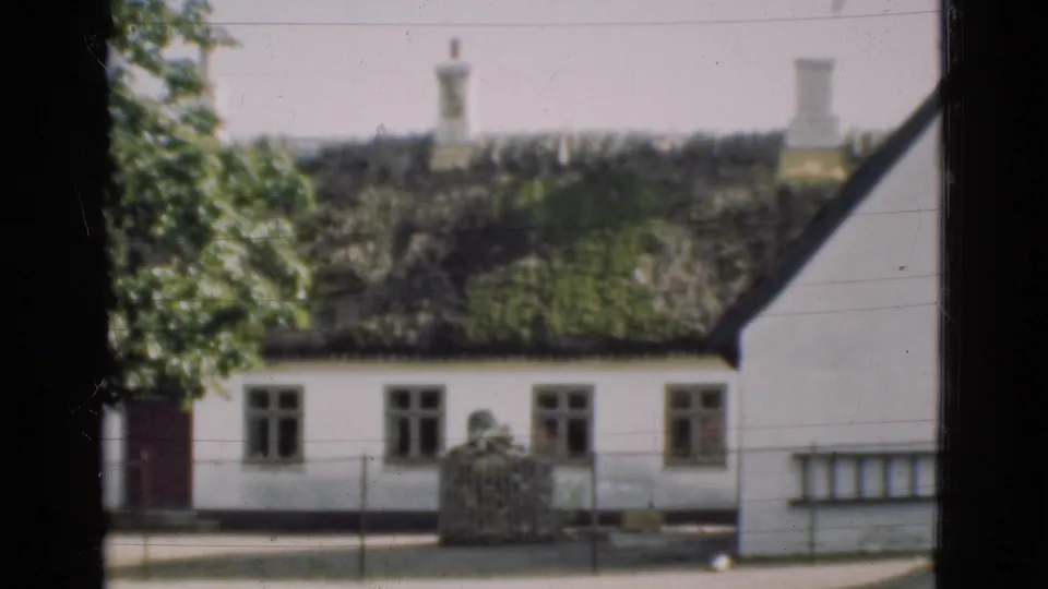 1975 Denmark frame with a white country house, mossy thatched roof, chimneys, tree leaves, roadside fence, and dark window edge.