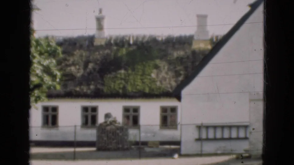 1975 Denmark home-movie view of the lower facade of a white thatched house with windows, fence, tree edge, and foreground framing.
