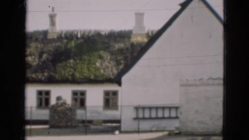 1975 Denmark frame showing a white country house wall, dark thatched roof, windows, roadside fence, stacked material, and a ladder leaning at right.