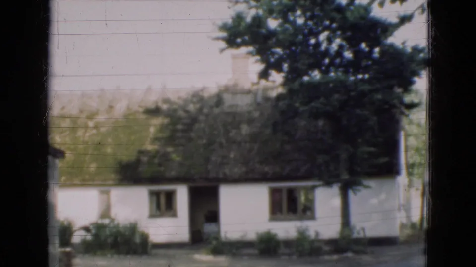 1975 Denmark 8mm view of a white thatched farmhouse, small windows, trees, shrubs, and dark window-frame borders.