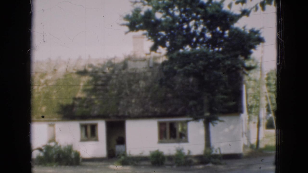 White farmhouse with thatched roof, open doorway, trees, and grass viewed from behind a dark frame.
