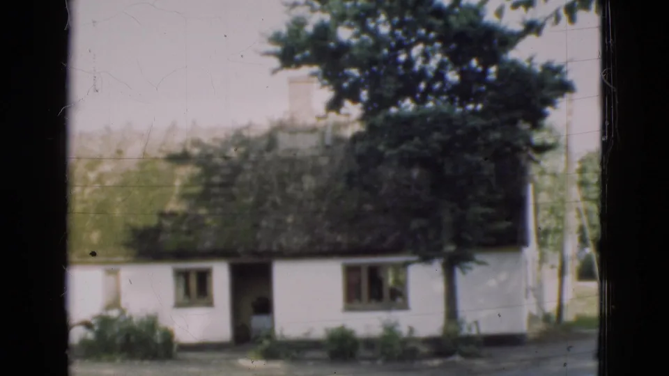 1975 Denmark home-movie frame of a white thatched farmhouse with trees, grass, windows, and a dark vertical frame edge.