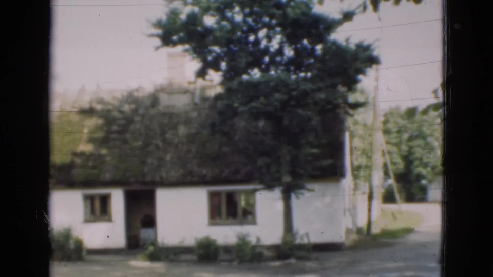 1975 Denmark 8mm frame showing a closer farmhouse exterior with thatched roof, open doorway, windows, trees, shrubs, and road edge.