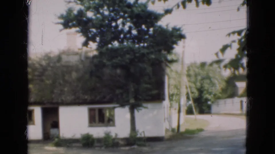 1975 Denmark home-movie view of a white thatched farmhouse by a road, with trees, shrubs, windows, and a possible figure in the doorway.