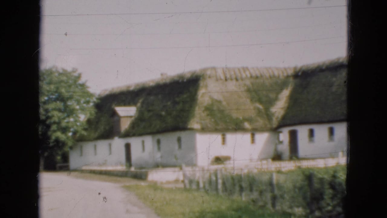 1975 Denmark 8mm frame showing a white rural farmhouse with a mossy thatched roof, dirt path, fence, and dark window edge.