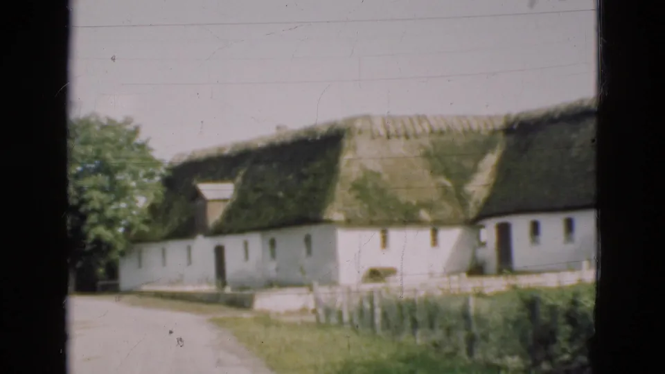 1975 Denmark 8mm frame showing a white rural farmhouse with a mossy thatched roof, dirt path, fence, and dark window edge.