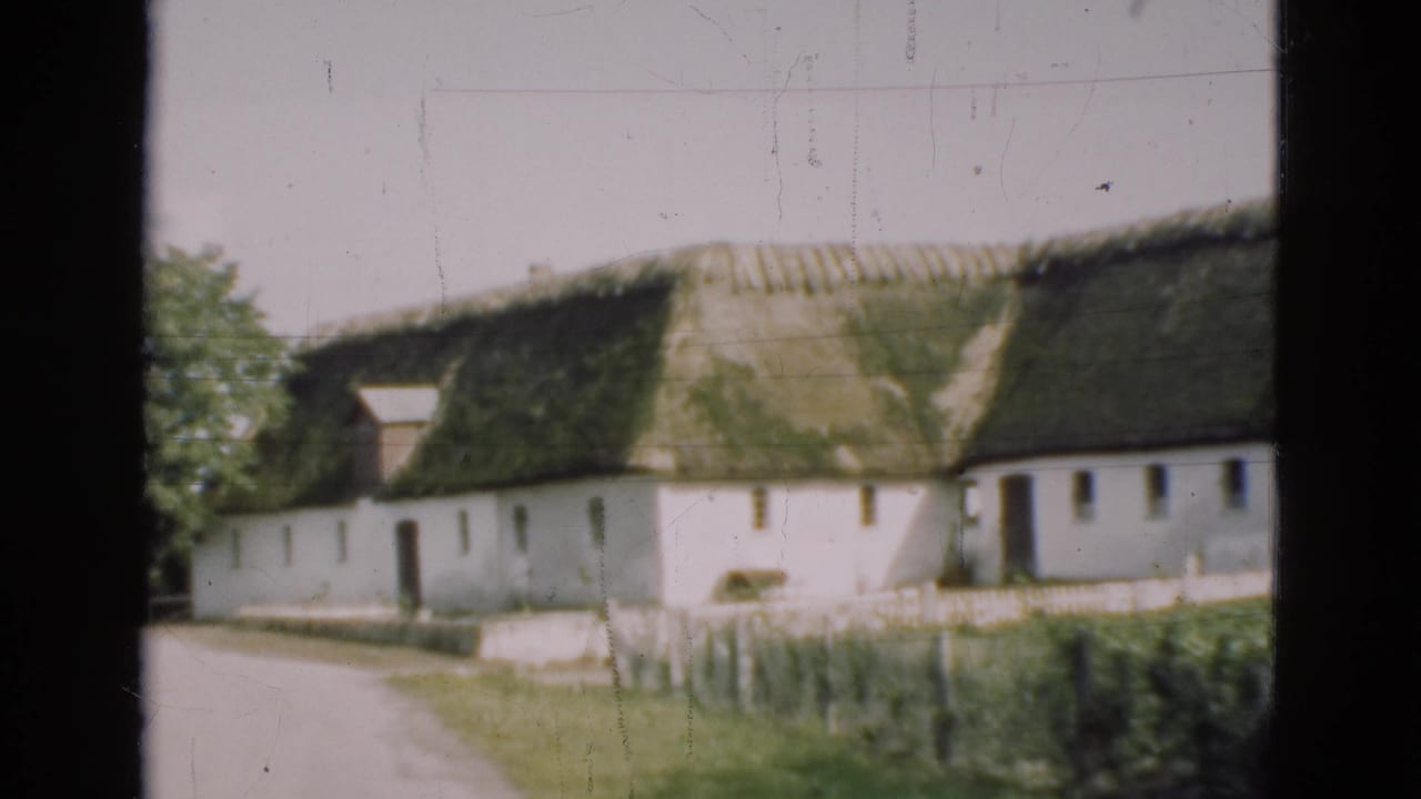 Thatched white farmhouse beside a path, partly framed by dark vertical window edges.