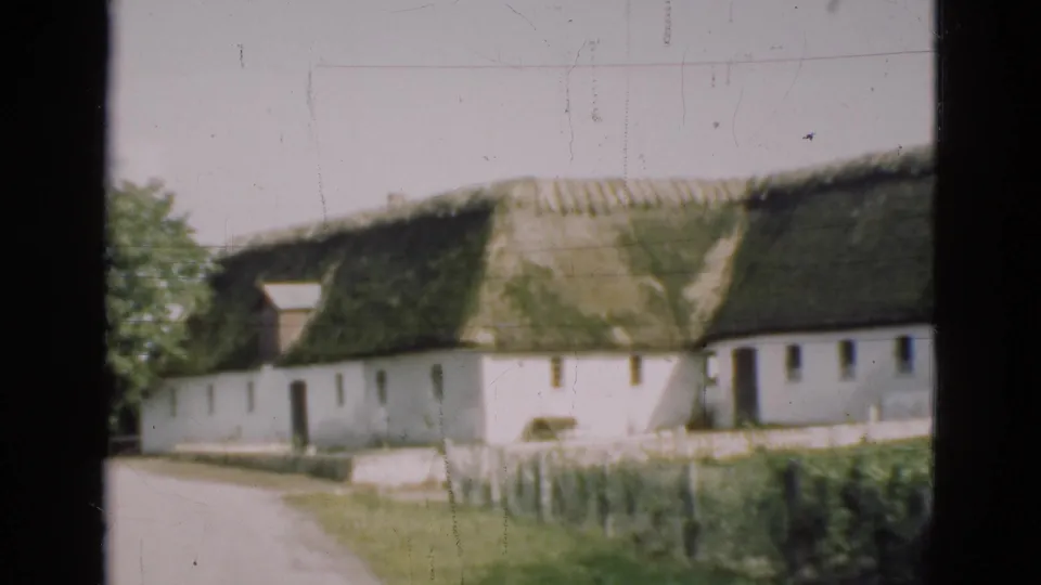 1975 Denmark home-movie frame with a white farmhouse, green thatched roof, grassy fence line, trees, and a dark interior frame edge.
