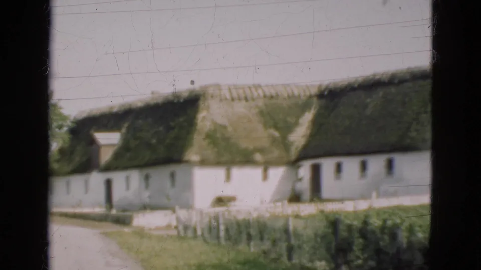 1975 Denmark frame showing the farmhouse closer in soft focus, with white walls, mossy thatch, path, fence posts, and grass.