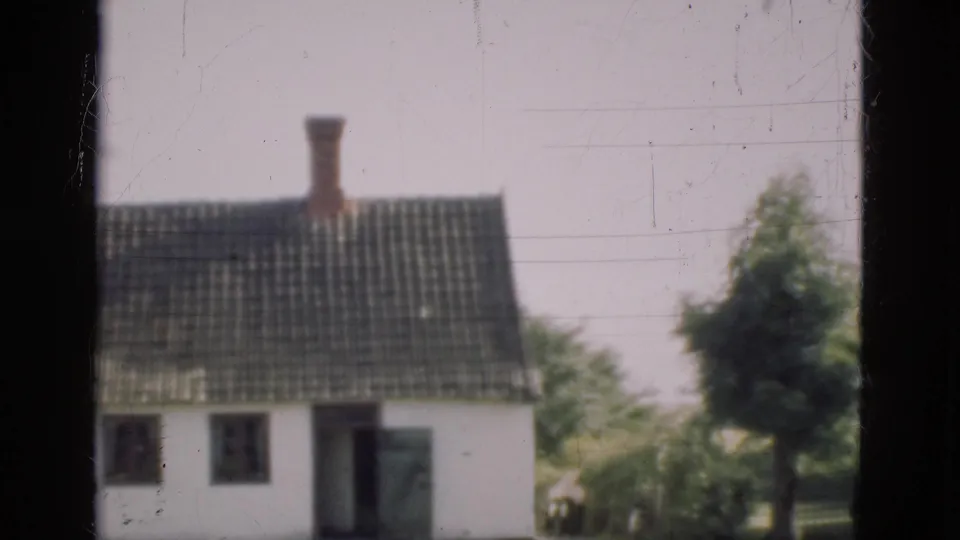 1975 Denmark 8mm frame of a white rural house with dark tiled roof, chimney, doorway, tree, and garden area in soft home-movie focus.