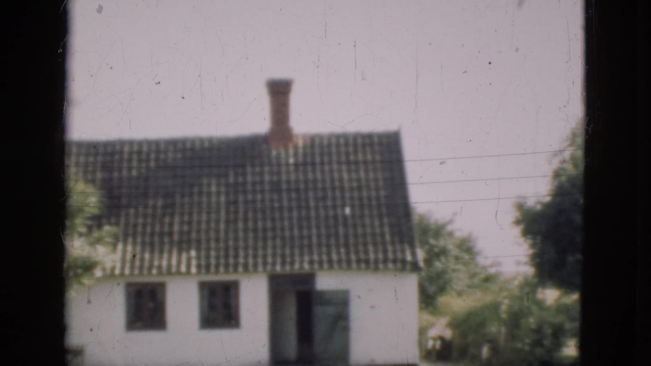 1975 Denmark 8mm frame showing a white house with a dark tiled roof and chimney viewed through dark window bars.