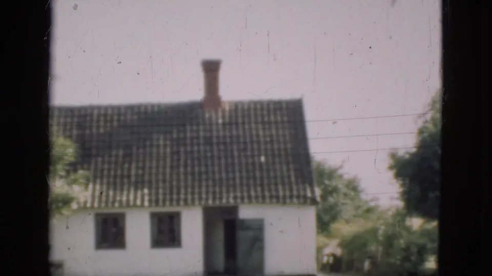 1975 Denmark 8mm frame showing a white house with a dark tiled roof and chimney viewed through dark window bars.